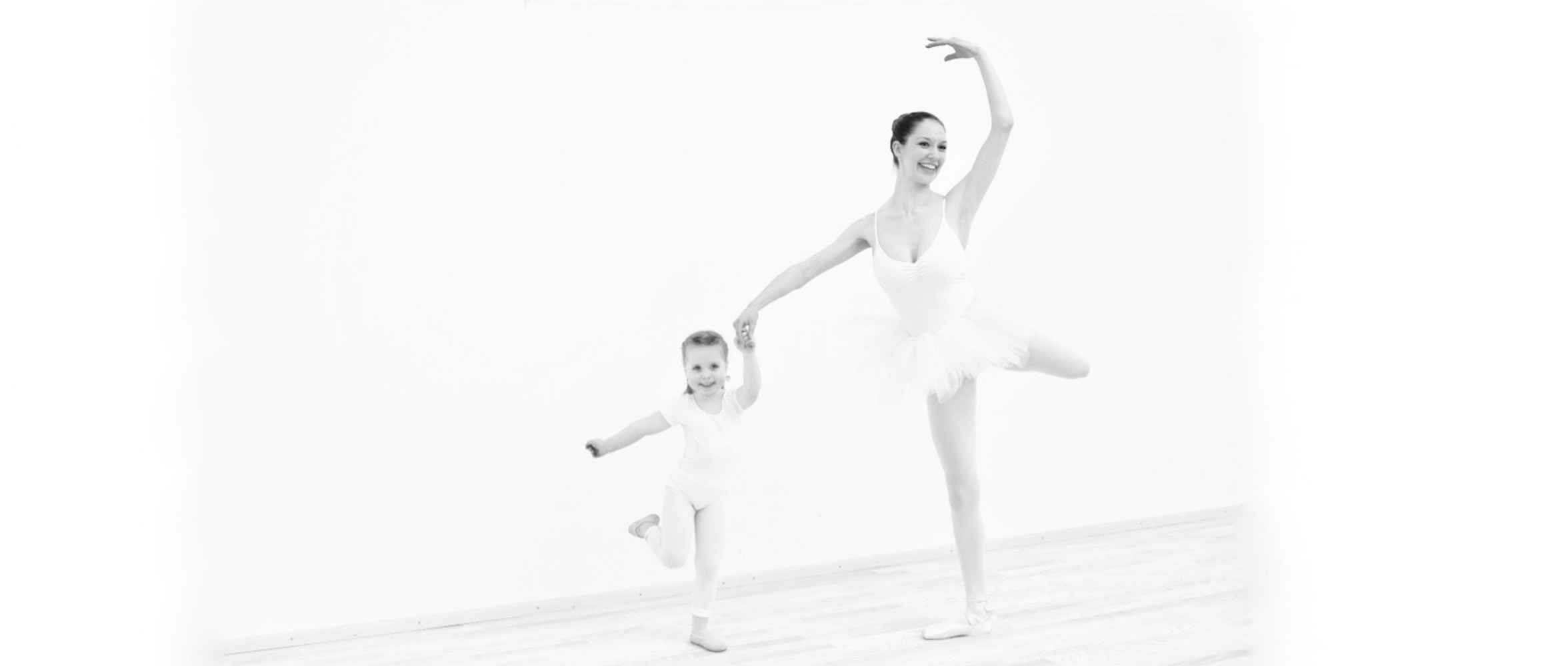 Erwachsene und junge Balletttänzerinnen stehen gemeinsam einbeinig in Pose im Ballettoutfit und strahlen in die Kamera
